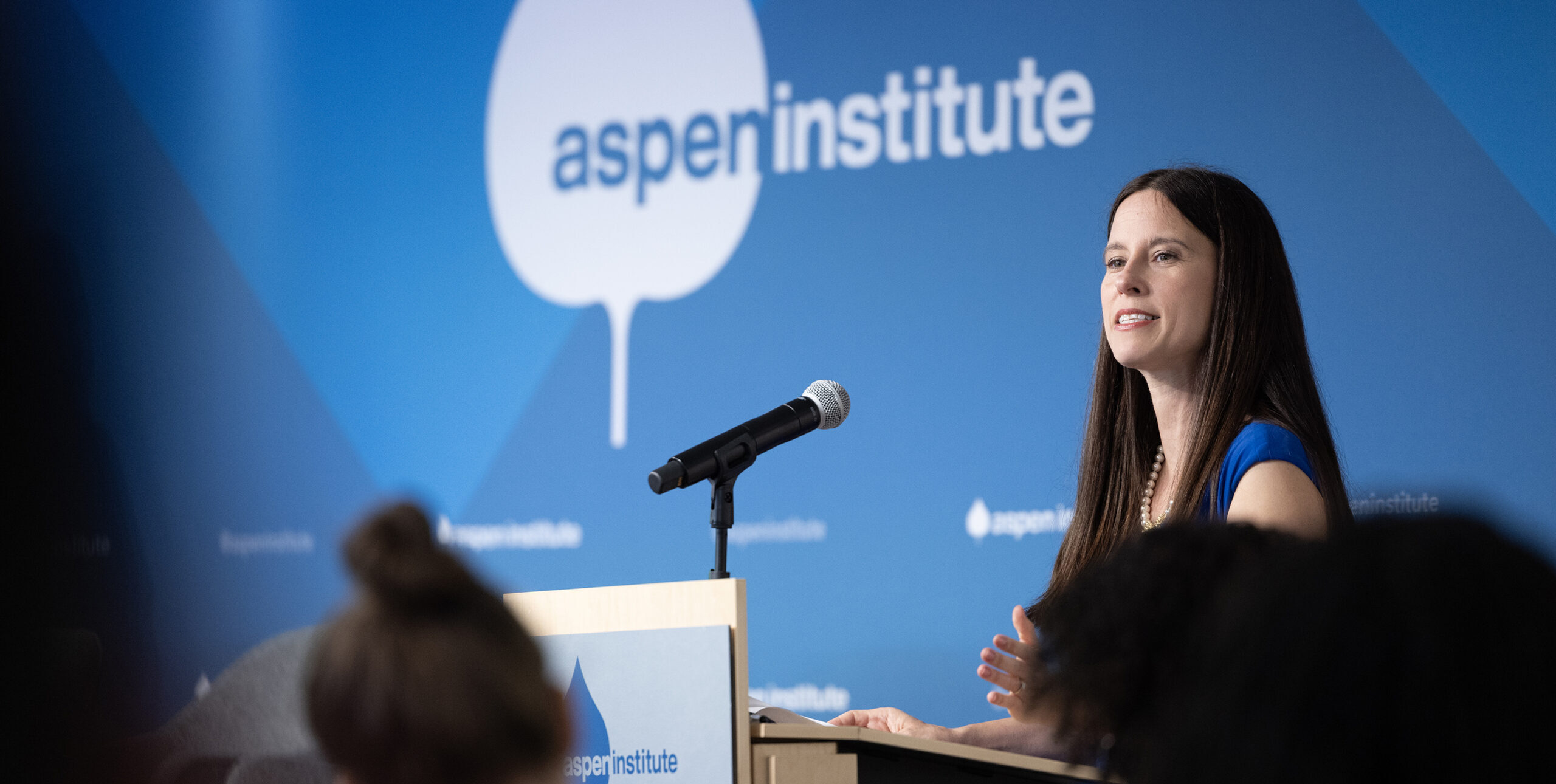 Academy Director Betsy Cooper gestures to the audience while speaking into a microphone at a podium. Behind her is a dark blue backdrop with the Aspen Institute's logo in white.