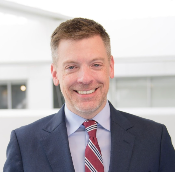 Ross Gillfillan smiles at the camera in a dark blue suit jacket, light blue collared shirt, and striped red and white tie.