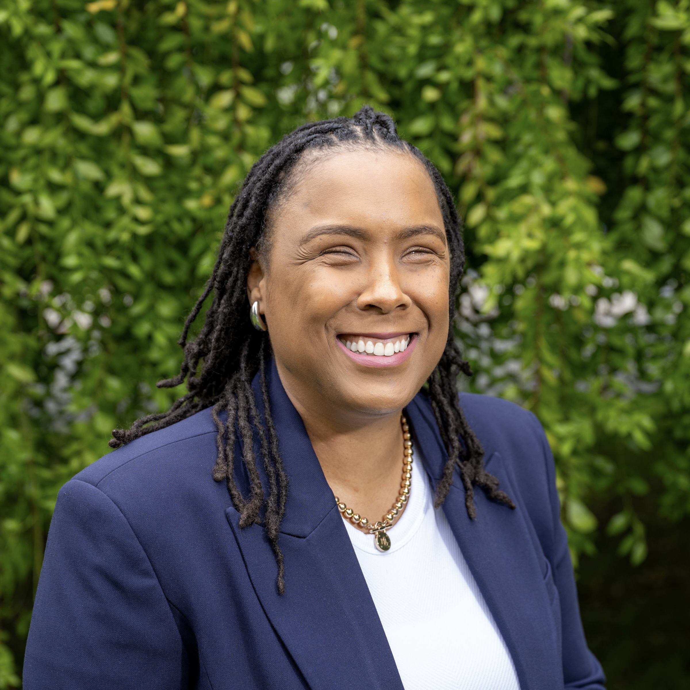 Tiffani smiles at the camera in a white blouse and dark blue suit jacket against a backdrop of hanging vines.