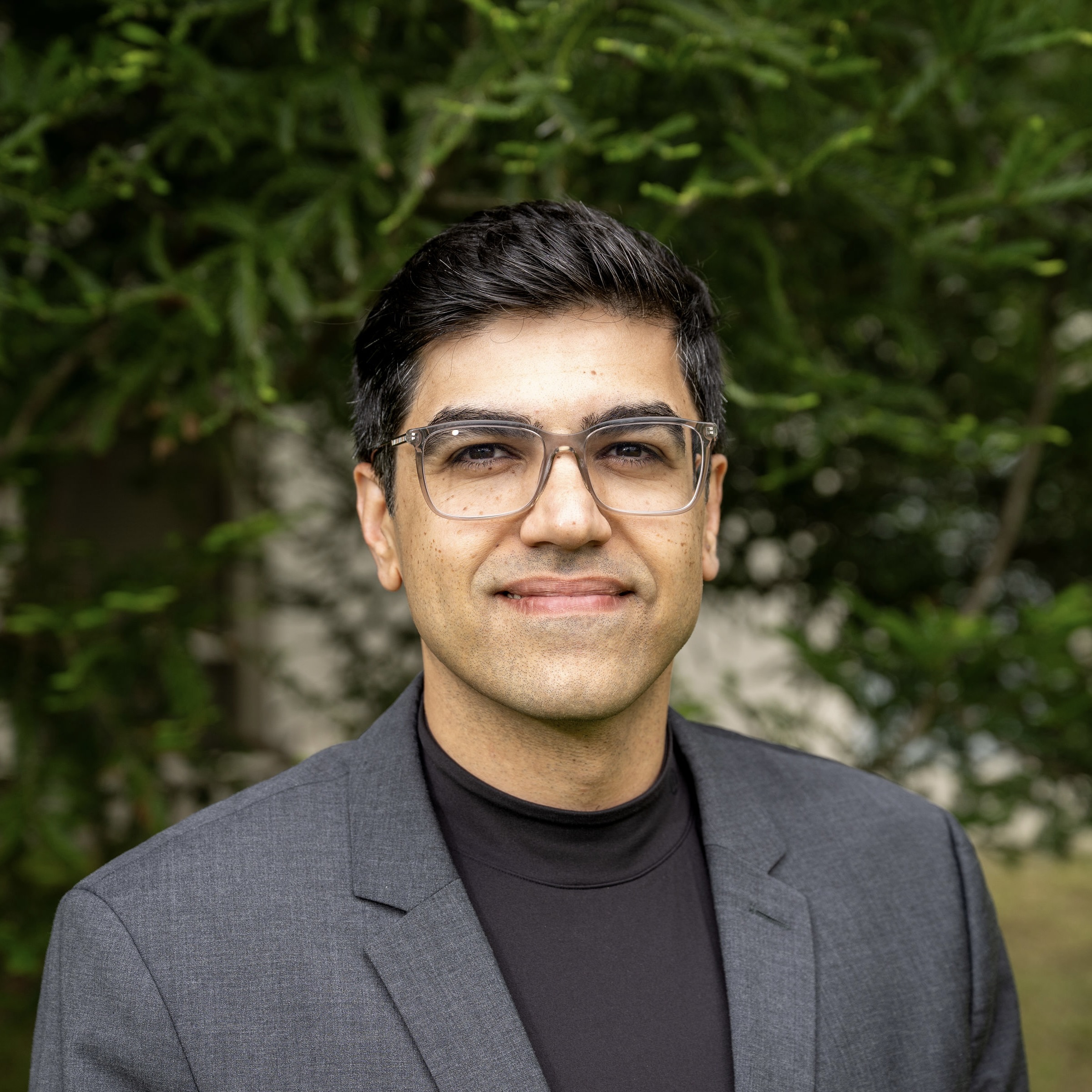 Amin smiles at the camera in a black shirt and dark grey suit jacket.