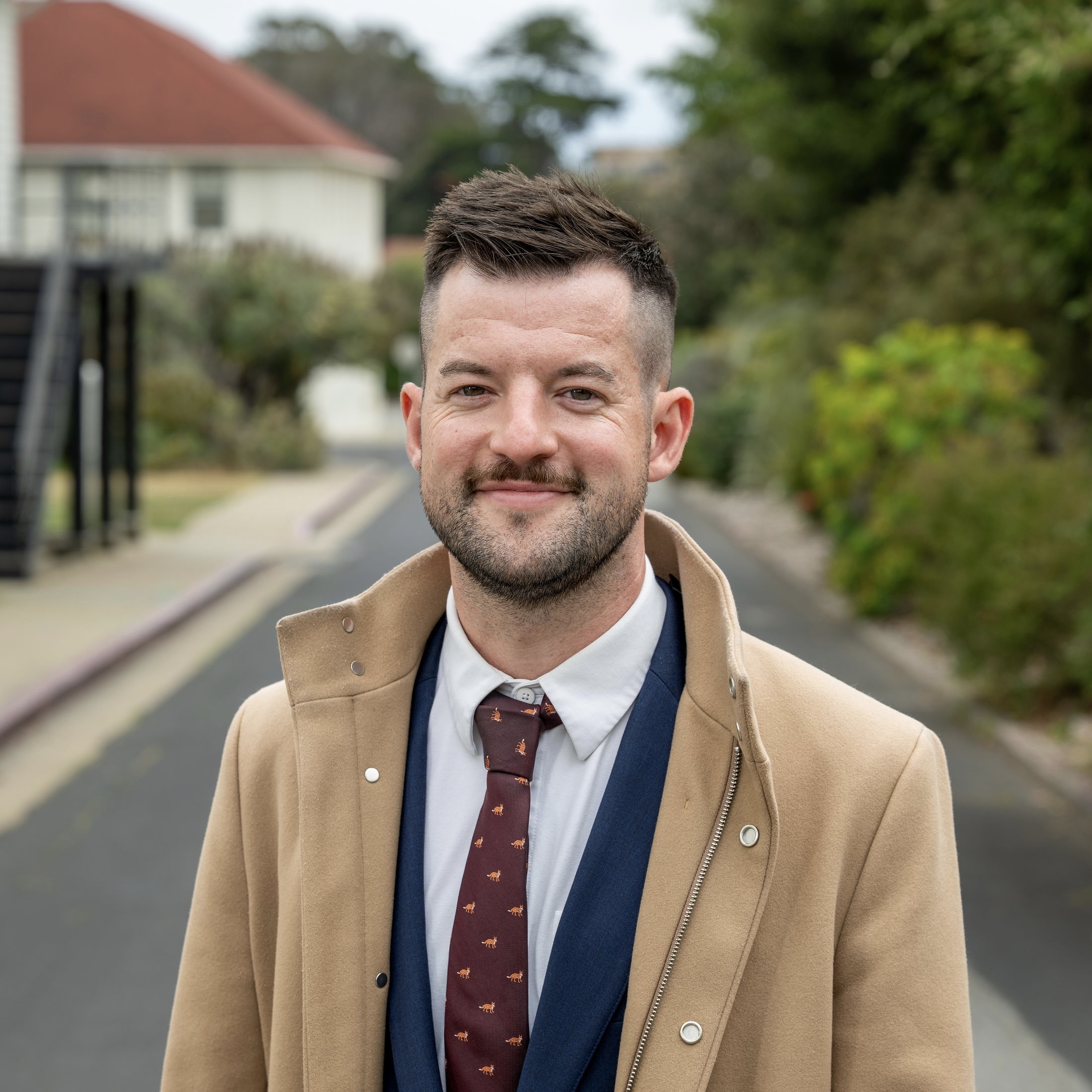 Will smiles at the camera in a white collared shirt, red tie, and tan trench coat.
