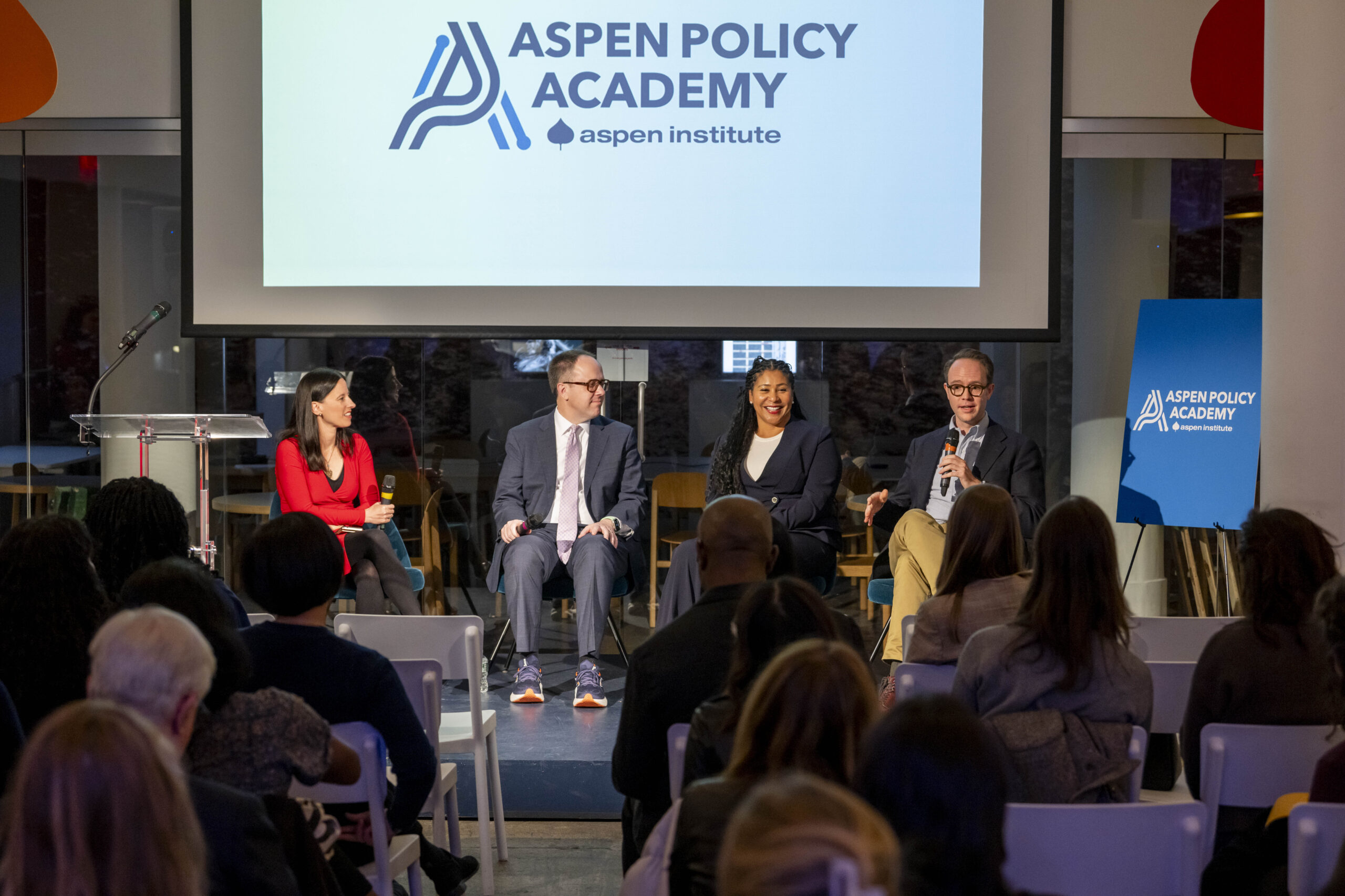 Aspen Policy Academy Director Betsy Cooper and another guest facilitate a panel with former Mayors London Breed and G.T. Bynum. The 4 speakers sit in chairs on stage with the Aspen Policy Academy logo displayed on a projector screen behind them.