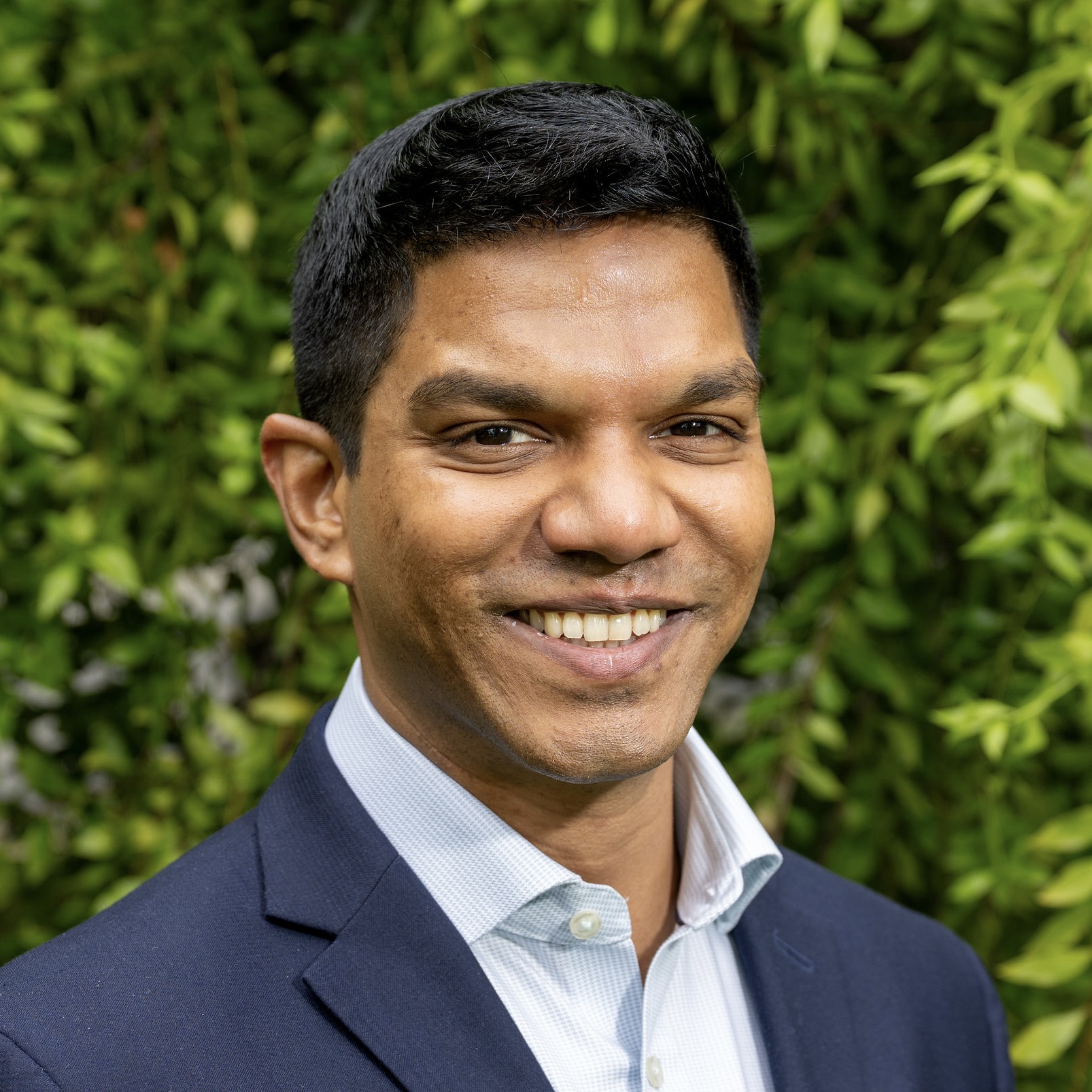 Roby smiles in a light blue collared shirt and dark blue suit jacket.