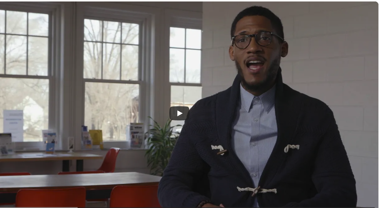 A screenshot of a YouTube video showing a man speaking to the camera in a classroom wearing a blue shirt and dark blue sweater.