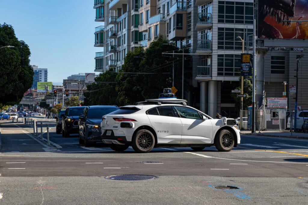 A white Waymo (an autonomous vehicle company) crosses the street at a busy, city-center intersection.