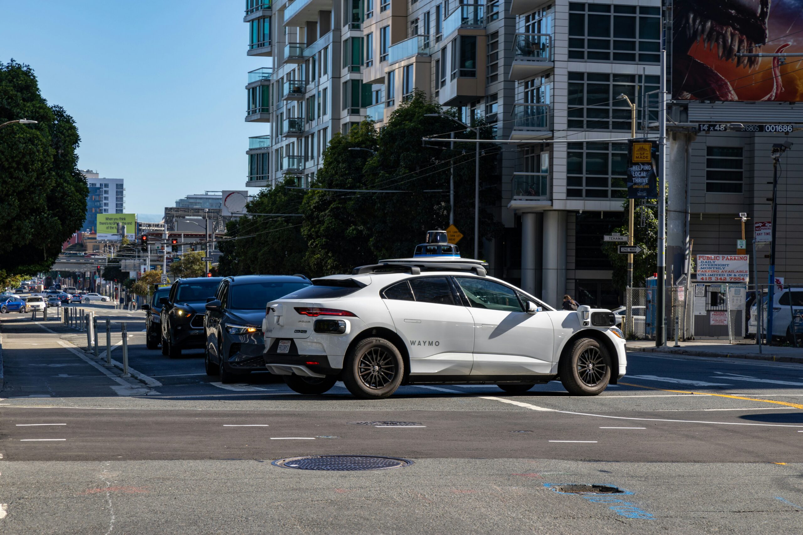 A white Waymo (an autonomous vehicle company) crosses the street at a busy, city-center intersection.