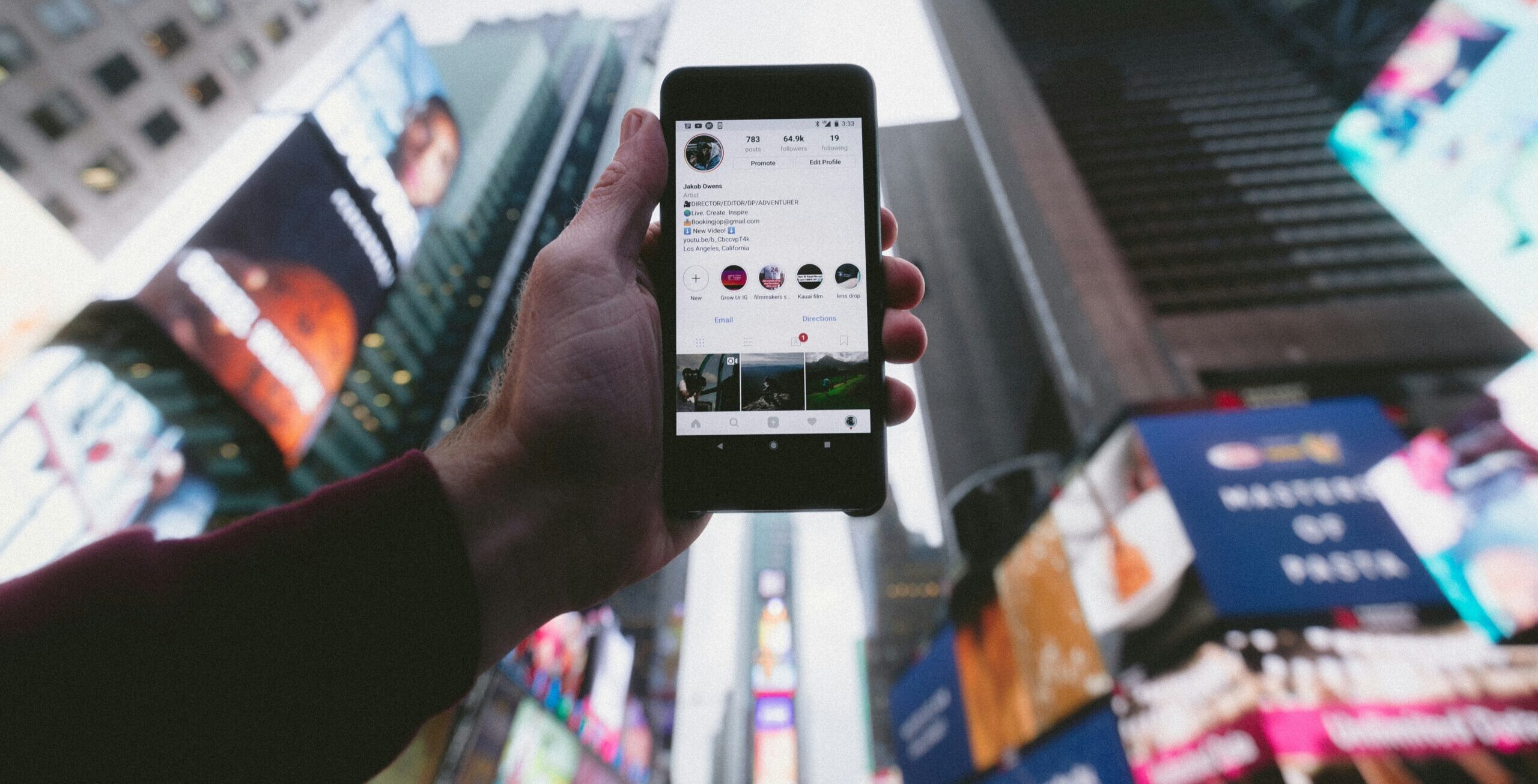 A person holds up a cell phone showing their Instagram account with tall city buildings in the background.