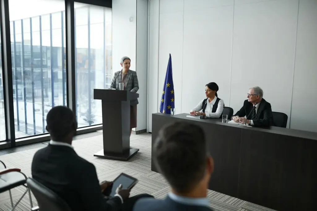 Woman standing at a podium speaking to government officials in a conference room.
